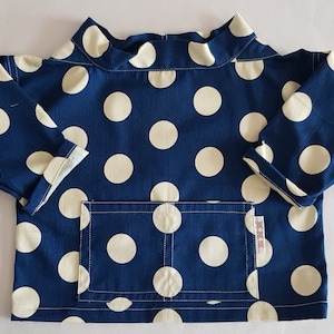 May include: A blue long-sleeved shirt with white polka dots and a patch pocket. The shirt has a high neckline and a relaxed fit.
