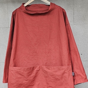 Könnte beinhalten: Ein rostfarbenes Langarmshirt mit hohem Ausschnitt und zwei großen Fronttaschen. Das Hemd hängt an einem Holzbügel vor grauem Hintergrund. Der Stoff scheint eine leichte Leinen- oder Baumwollmischung zu sein.