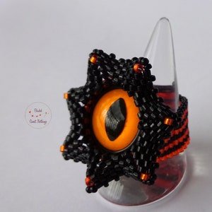 Könnte beinhalten: Ein schwarzer und orangefarbener Perlenring mit sternförmigem Design und einem großen orangefarbenen Auge in der Mitte. Der Ring befindet sich auf einem klaren Ringständer.