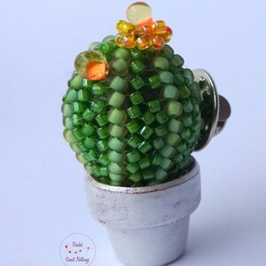 May include: A green beaded cactus in a white pot with a pin back. The cactus has orange and yellow beads for flowers.