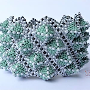 Puede incluir: Un brazalete de cuentas con un diseño geométrico en tonos verdes, plateados y negros. El brazalete está hecho con cuentas pequeñas, tejidas apretadamente y tiene un aspecto texturizado único.