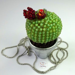 Puede incluir: Un collar de cactus de cuentas con flores rojas y una cadena plateada. El cactus está en una maceta plateada con tierra marrón.