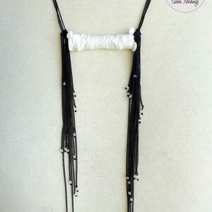 May include: A white and black beaded necklace with a long, rectangular pendant. The pendant is made of white fabric and has a woven texture. The necklace is on a white mannequin.