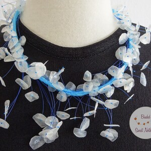 May include: A blue and white necklace made of small, white, round, shell-like beads. The beads are strung on blue cord and hang in a cascading pattern. The necklace is on a black shirt. The text "Beaded Sweet Nothings" is in the lower right corner of the image.