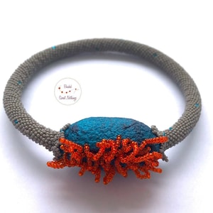 Może przedstawiać: Ręcznie robiony naszyjnik z szarą opaską z koralików i centralnym elementem z turkusowego filcu. Filc jest ozdobiony pomarańczowymi akcentami z koralików. Unikalna biżuteria.
