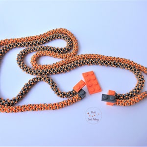 Peut inclure: Un long collier composé de perles grises et orange. Le collier est composé de deux brins de perles qui sont torsadés ensemble. Il y a trois petits blocs Lego gris et orange à côté du collier.