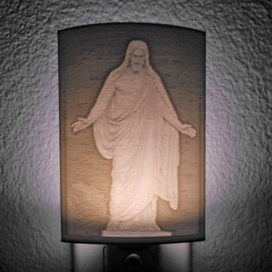 Può includere: Una luce notturna bianca con finitura in legno presenta un'immagine in rilievo di Gesù Cristo. La luce è accesa, proiettando un bagliore morbido sull'immagine.