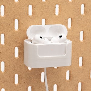 Könnte beinhalten: Weiße AirPods Pro-Ohrhörer in einem offenen Ladecase, montiert auf einer Lochwand. Das Case wird in einem weißen, 3D-gedruckten Halter gehalten. Ein weißes Ladekabel ist mit dem Case verbunden. Die Lochwand besteht aus hellbraunem Holz.