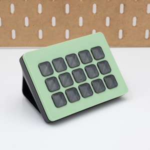 以下が含まれることがあります： ミントグリーンと黒の電子デバイス。16個の灰色の正方形のボタンが付いています。デバイスは上向きに傾斜しており、黒いベースが見えます。背景は明るい茶色のペグボードです。
