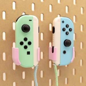 Könnte beinhalten: Zwei pastellfarbene Gamecontroller, einer mintgrün und der andere hellblau, sind an einer Lochwand befestigt. Jeder Controller wird von einem rosa Halter gehalten. Die Controller haben schwarze Knöpfe und Joysticks, mit einem Kabelanschluss.