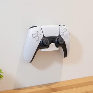 Può includere: Un controller bianco e nero per PlayStation 5 montato su una parete bianca. Il controller è tenuto da un supporto bianco. Il controller ha un design simmetrico con pulsanti e joystick. Una piccola pianta verde in un vaso grigio si trova nell'angolo in basso a sinistra.
