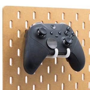 Puede incluir: Un mando negro de Xbox está montado en un tablero perforado marrón con un soporte blanco. El tablero perforado tiene una serie de ranuras verticales espaciadas uniformemente. El mando tiene varios botones y joysticks.