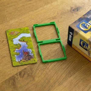 May include: A green plastic game piece holder next to a game card featuring a river and landscape design. The game card is rectangular with a colourful illustration. A game box is partially visible.