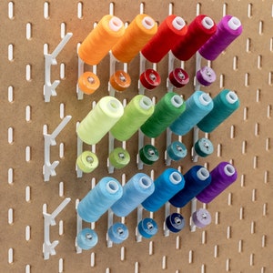 May include: A pegboard with white thread spool holders, each holding a spool of colorful thread. The thread colors include orange, red, purple, green, blue, and teal. Below each spool is a matching bobbin. The pegboard is a light brown color.