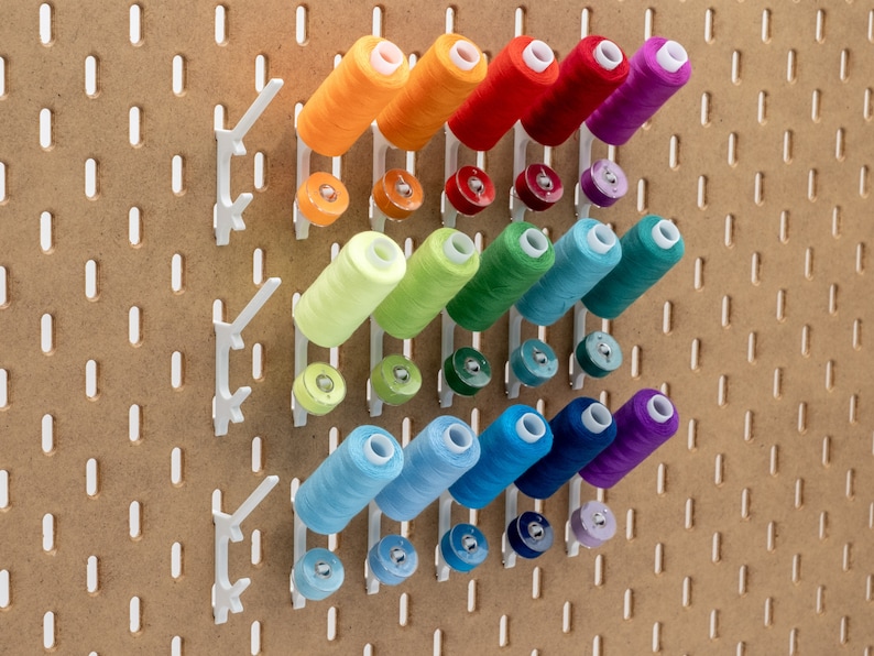 May include: A pegboard with white thread spool holders, each holding a spool of colorful thread. The thread colors include orange, red, purple, green, blue, and teal. Below each spool is a matching bobbin. The pegboard is a light brown color.