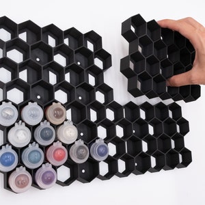 Op de afbeelding: Zwart modulair verfrek met een honingraatontwerp, met diverse verfpotjes. Het rek is ontworpen om uit te breiden en wordt getoond met een hand die een apart gedeelte vasthoudt. De verfpotjes zijn gevuld met verschillende kleuren.