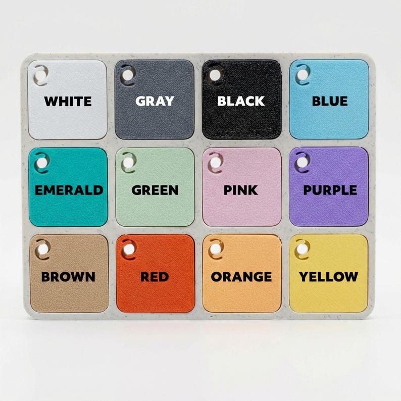 May include: A colour palette featuring twelve square colour swatches in a rectangular tray. Colours include white, grey, black, blue, emerald, green, pink, purple, brown, red, orange, and yellow. Each swatch has a hole and a colour name.