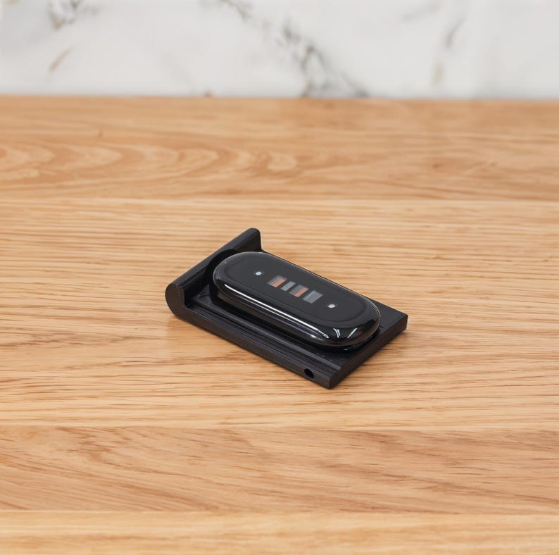Puede incluir: Una base de carga de pl&aacute;stico negro para un monitor de actividad f&iacute;sica, con una base rectangular de borde elevado y un conector magn&eacute;tico ovalado central. El dispositivo es negro brillante con una peque&ntilde;a pantalla que muestra cuatro barras de color.