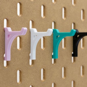 Puede incluir: Cuatro ganchos de tablero perforado de colores, rosa, blanco, turquesa y negro, insertados en un tablero perforado marrón. Los ganchos son de plástico y tienen un diseño curvo para sujetar objetos. El tablero perforado tiene agujeros espaciados.