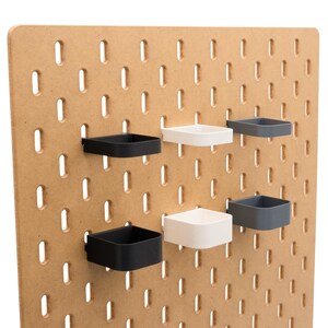 May include: A light brown pegboard with evenly spaced oval holes. Attached are six square, open-topped containers in black, white, and gray. The containers are designed to hold small items and are mounted on the pegboard.