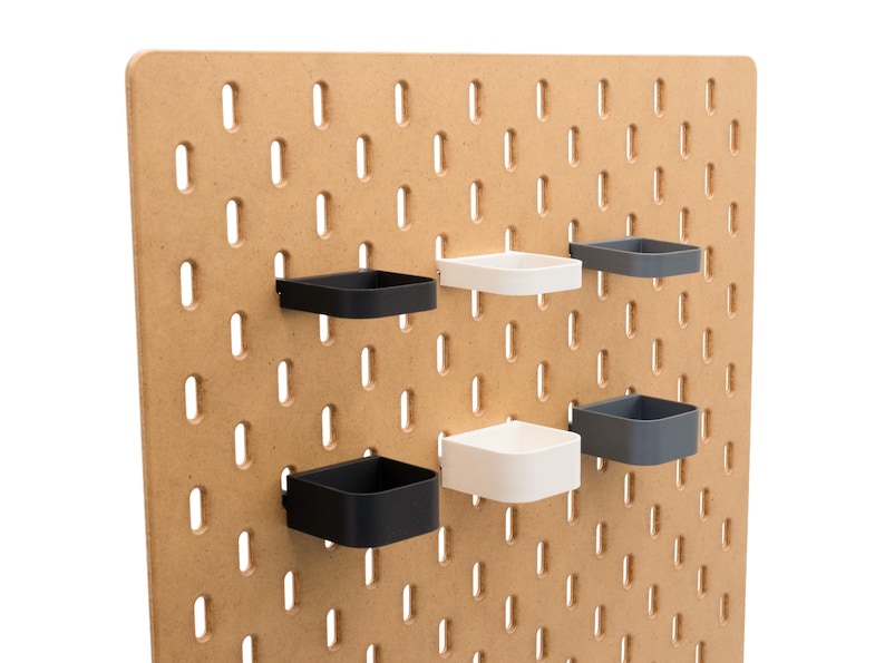 May include: A light brown pegboard with evenly spaced oval holes. Attached are six square, open-topped containers in black, white, and gray. The containers are designed to hold small items and are mounted on the pegboard.