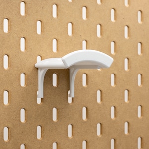 May include: A white pegboard shelf fixed to a brown pegboard. The shelf features a curved top and is designed for storage. The pegboard has evenly spaced oval holes.