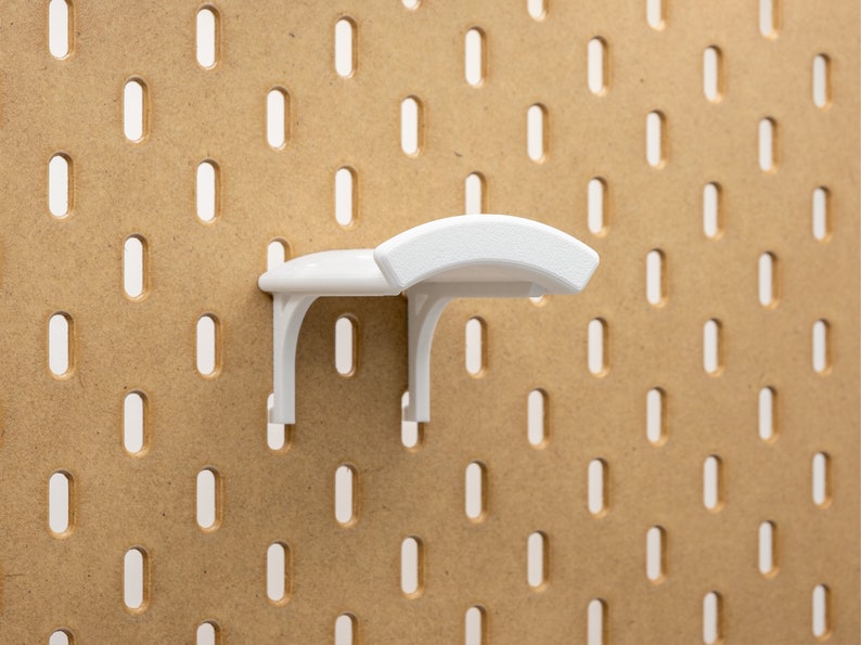 May include: A white pegboard shelf fixed to a brown pegboard. The shelf features a curved top and is designed for storage. The pegboard has evenly spaced oval holes.