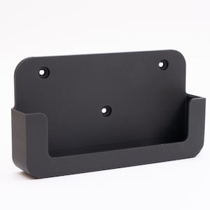Puede incluir: Un soporte de pared negro mate con un diseño rectangular. Cuenta con un pequeño estante y tres orificios pretaladrados para el montaje. El soporte está hecho de un material sólido, posiblemente impreso en 3D.