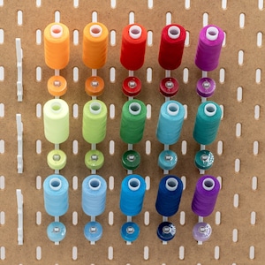 May include: A collection of colorful spools of thread and bobbins arranged on a pegboard. The thread colors include orange, red, green, blue, and purple. The bobbins are in matching colors. The pegboard is made of light brown wood with white plastic pegs.