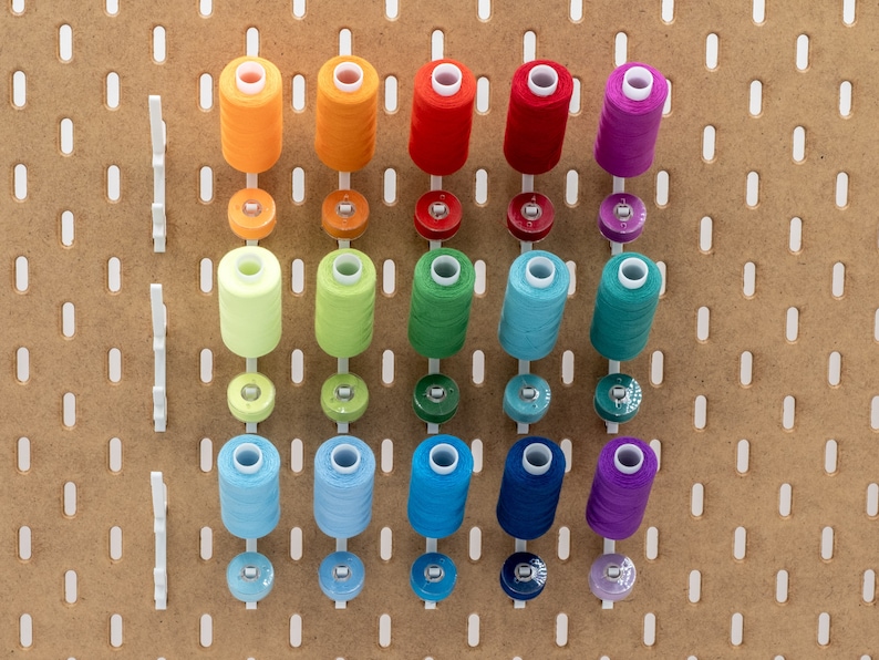 May include: A collection of colorful spools of thread and bobbins arranged on a pegboard. The thread colors include orange, red, green, blue, and purple. The bobbins are in matching colors. The pegboard is made of light brown wood with white plastic pegs.