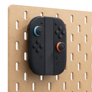 May include: A black games controller with blue and orange detailing is mounted on a light brown pegboard. The controller features buttons and joysticks. The pegboard has evenly spaced rectangular holes.