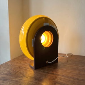 May include: A retro table lamp featuring a bright yellow, semi-circular shade and a dark brown, arched base. The lamp emits a warm light. The white cord is visible. The lamp is on a wooden surface.