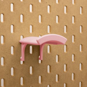 May include: A light pink pegboard shelf accessory. The shelf has a curved top and two supporting legs. It is mounted on a brown pegboard with evenly spaced oval holes.