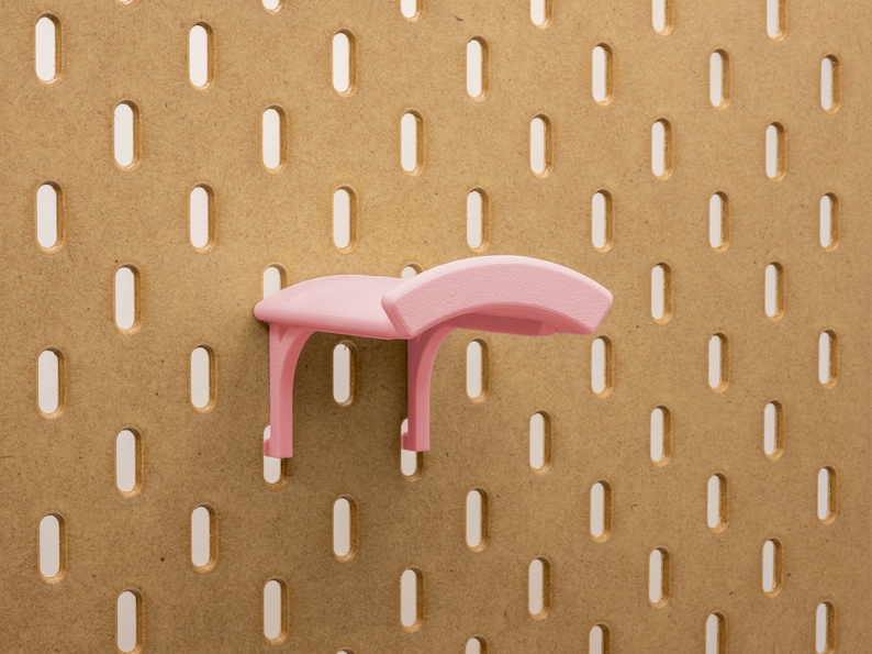 May include: A light pink pegboard shelf accessory. The shelf has a curved top and two supporting legs. It is mounted on a brown pegboard with evenly spaced oval holes.