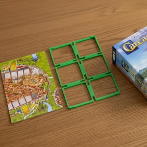 Puede incluir: Un juego de mesa Carcassonne con una tarjeta de juego que muestra un mapa detallado de una ciudad medieval, soportes de fichas de plástico verde y la caja del juego. La tarjeta del juego presenta un río, edificios y carreteras.