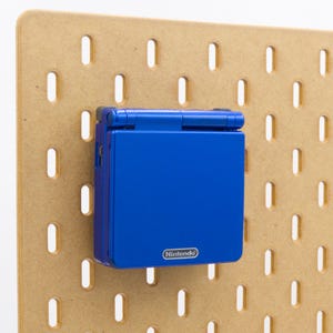 Può includere: Una console di gioco portatile Nintendo Game Boy Advance SP blu attaccata a una pannello forato marrone chiaro. La console è chiusa e mostra il logo Nintendo.