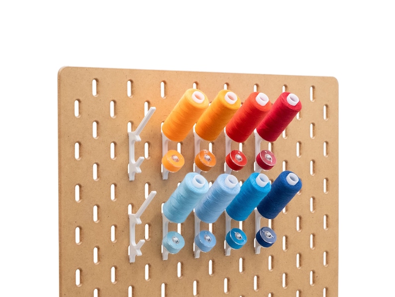 May include: A wooden pegboard with white plastic holders displaying spools of thread in various colors. The thread colors include orange, red, and shades of blue. The pegboard is designed for organizing sewing supplies.