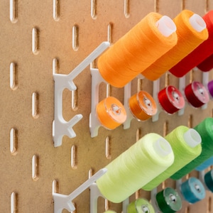 May include: A wooden pegboard with white plastic holders displaying colorful spools of thread and bobbins. The thread colors include orange, red, purple, green, and blue. The pegboard has evenly spaced holes.