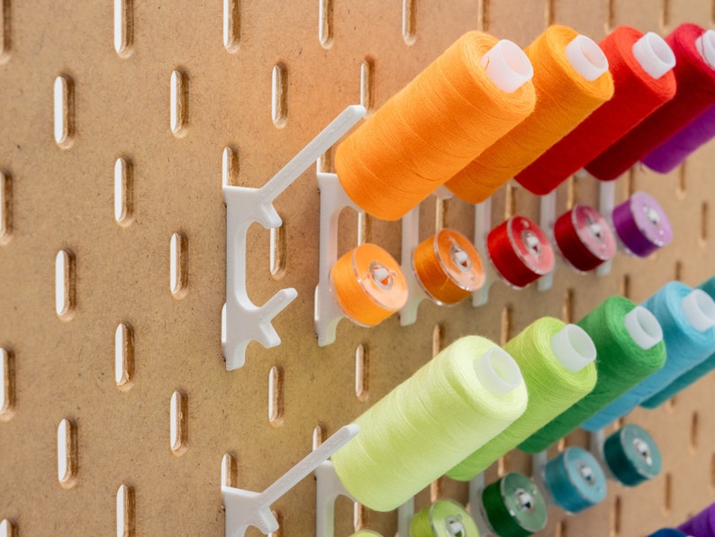 May include: A wooden pegboard with white plastic holders displaying colorful spools of thread and bobbins. The thread colors include orange, red, purple, green, and blue. The pegboard has evenly spaced holes.