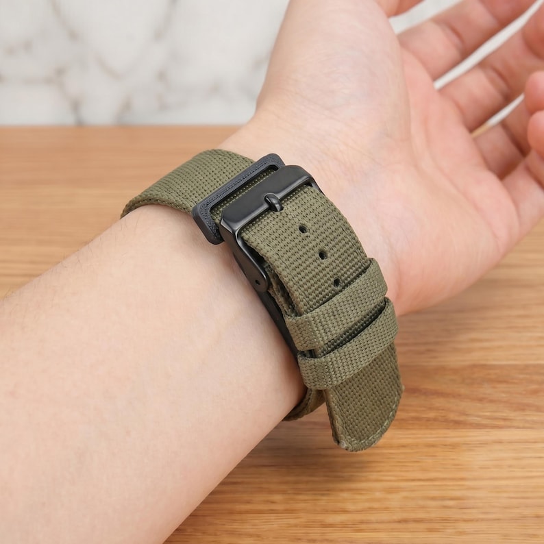 K&ouml;nnte beinhalten: Ein olivgr&uuml;nes Uhrenarmband aus Canvas mit einer mattschwarzen Metallschnalle und einer passenden schwarzen Metallschlaufe. Das strukturierte Stoffband verf&uuml;gt &uuml;ber eine Reihe vorgestanzter L&ouml;cher zur Anpassung und einen stabilen Dornschlie&szlig;enverschluss.