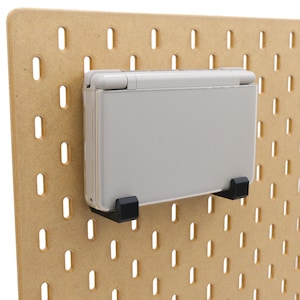 May include: A light grey handheld games console mounted on a pegboard with black brackets. The pegboard is made of light brown wood and has evenly spaced oval holes.