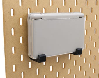 Op de afbeelding: Een lichtgrijze handheld gameconsole gemonteerd op een pegboard met zwarte beugels. Het pegboard is gemaakt van lichtbruin hout en heeft gelijkmatig verdeelde ovale gaten.