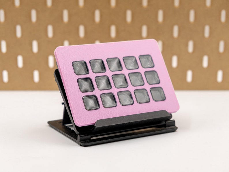 May include: A pink, rectangular device with sixteen square, dark gray buttons. The device is mounted on a black, adjustable stand. The background is a brown pegboard with white, vertical cutouts.