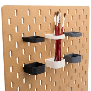 May include: A wooden pegboard with multiple small, square containers in black, white, and gray. Red paint brushes are stored in a white container. The pegboard has evenly spaced oval holes.