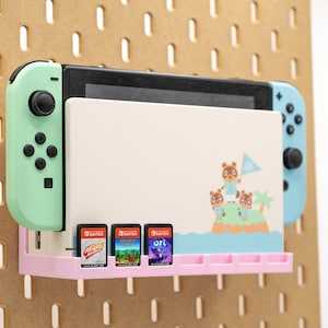 Puede incluir: Una consola Nintendo Switch con Joy-Cons verde menta y azul claro está montada en una panel perforado. Una estantería rosa contiene tres cartuchos de juego. La consola presenta un diseño de Animal Crossing.