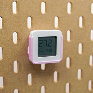 Può includere: Un piccolo termometro digitale quadrato, con un quadrante bianco e una cornice rosa, è montato su una pannello forato marrone. Il display mostra la temperatura a 27,2 gradi Celsius, con un intervallo di (-~148).