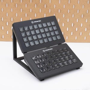 May include: Black Stream Deck with multiple buttons, resting on a black stand. The Stream Deck has the words "Stream Deck" printed on it. The background is a light brown pegboard.