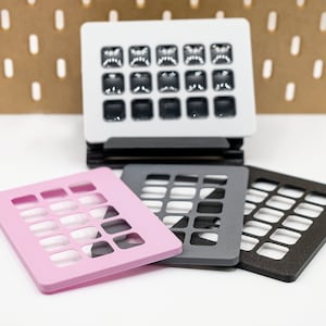 May include: A stack of black coasters supports a white square object with multiple small, dark squares. Three additional coasters in pink, gray, and brown are in the foreground. The background features a pegboard.