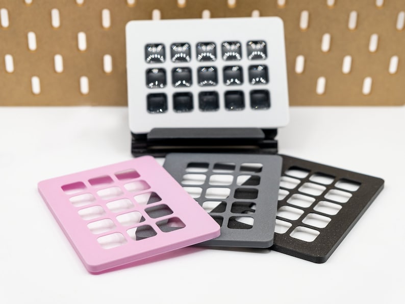 May include: A stack of black coasters supports a white square object with multiple small, dark squares. Three additional coasters in pink, gray, and brown are in the foreground. The background features a pegboard.