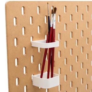May include: A light brown pegboard with evenly spaced holes. Two white, square-shaped holders are attached, each holding several red paint brushes. The brushes have dark brown bristles and are of varying sizes.
